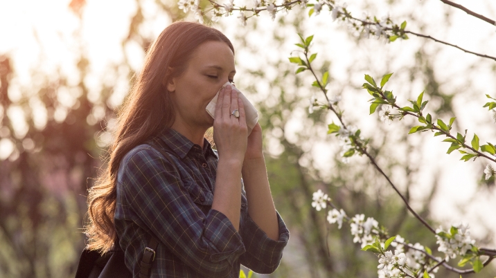 Frau mit Heuschnupfen-Symptomen putzt sich umgeben von Blüten die laufende Nase.